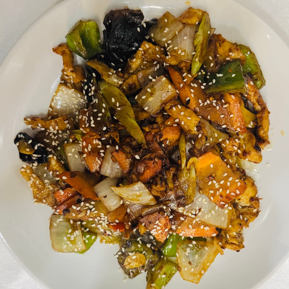 Stir-fried mixed vegetables, including peppers, onions, carrots, eggplant, and mushrooms, garnished with sesame seeds on a white plate.