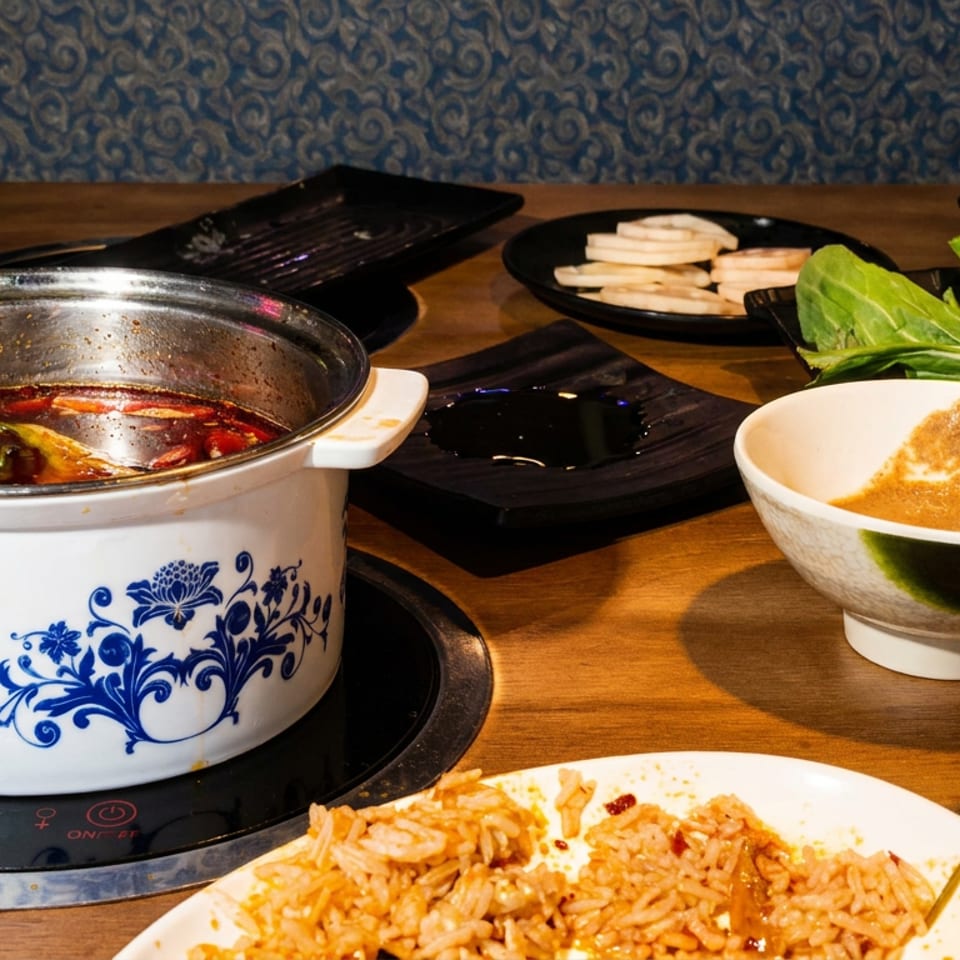 Chinese hot pot with spicy broth, dipping sauce, fresh vegetables, sliced ingredients, and fried rice on a restaurant table.